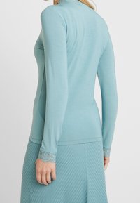 Long-sleeve top in light teal fabric with a smooth texture. Features lace accents at the cuffs, complementing a matching patterned skirt.