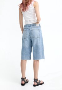 Shorts vaqueros en azul claro con dobladillo deshilachado. Vista trasera muestra dos bolsillos y una etiqueta de cuero con la marca. Combinados con sandalias negras de tiras.