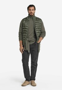 Grön pufferväst över en grön tröja, kombinerad med mörkgrå byxor och beige skor. Vesten har horisontell quiltning och en dragkedja fram.