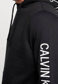 Černá mikina s kapucí, na rukávu bílý nápis "CALVIN". Materiál působí jemně a hladce. Součástí jsou šňůrky na stažení.