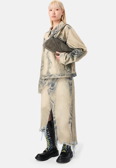 Donna che indossa una giacca in denim beige acid-wash e una gonna con orlo sfrangiato, mentre tiene una pochette in suede nera e stivali neri stringati con lacci colorati.