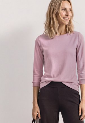 Lächelnde Frau mit schulterlangem blondem Haar trägt ein pinkfarbenes Langarmoberteil und schwarze Hose, sie hält eine kleine braune Tasche in ihrer rechten Hand.