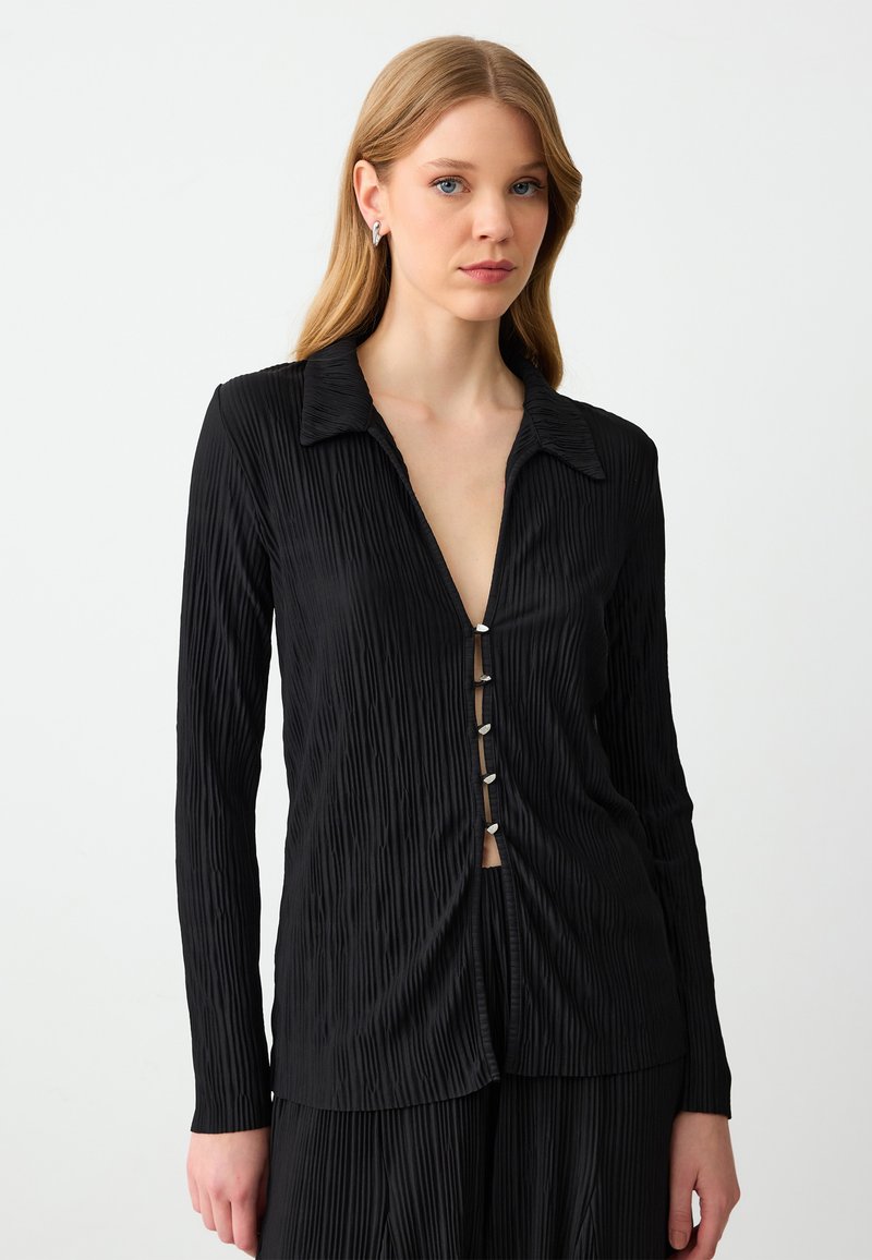 Zwarte, langemouwen shirt met een plissé textuur, puntige kraag en vijf decoratieve knopen aan de voorkant, wat een subtiele V-halslijn creëert.