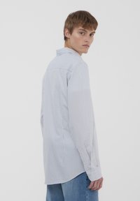 Camicia a righe blu chiaro con colletto classico, maniche lunghe e orlo leggermente curvato. Il tessuto appare liscio con dettagli a bottoni.