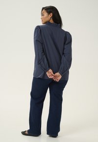 Cămașă albastră marin cu mâneci lungi și umăr rotunjit, cu guler plisat și textură netedă, purtată cu pantaloni flared din denim închis și pantofi negri.