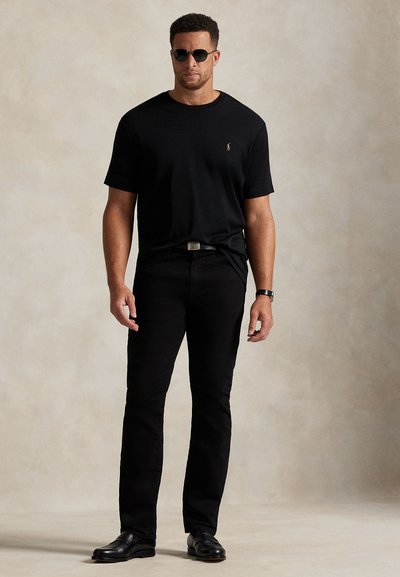 Mann iført svart kortermet skjorte, svarte bukser, svarte skinnsko, solbriller og armbåndsur står mot en nøytral bakgrunn.