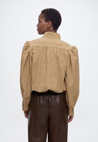 Γυναίκα με πλάτη φοράει μπλούζα από καφέ σουέτ με φουσκωτά μανίκια και ψηλόμεσο σκούρο καφέ δερμάτινο παντελόνι μπροστά από μονόχρωμο φόντο.