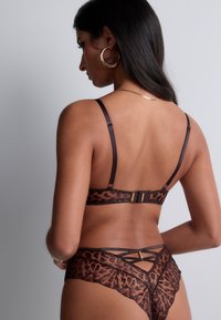 Ensemble de lingerie en dentelle brune présentant un motif léopard, des bretelles ajustables et des accents en métal doré. La vue de dos met en avant un design entrecroisé.