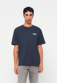 Joven con cabello corto y oscuro, vistiendo una camiseta azul marino de "Hike Club" y jeans gris claro, de pie contra un fondo blanco.