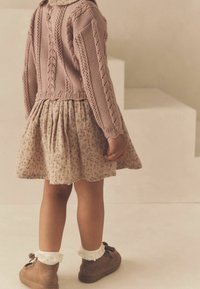 Pull en tricot rose avec un motif texturé, robe beige florale avec des plis, chaussettes en dentelle crème, et chaussures marron avec des nœuds.