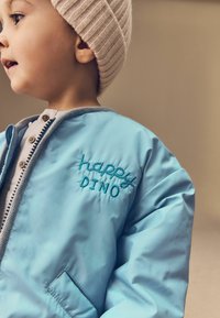 Ljusblå bomberjacka med inbroderad text "happy DINO", grå kant och en beige ribbad stickad mössa. Mjuk, glänsande tyg, framficka synlig.