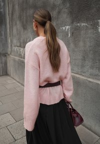 Rozā adīta džemperis ar ribotu apakšmalu, apvienots ar melnu plisētu svārku un bordo roku somu, uz teksturētas pelēkas sienas fona.