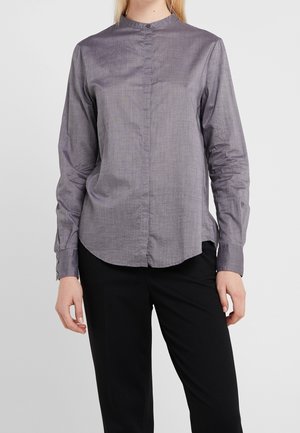 Person iført en langærmet, grå skjorte med knapper og kravebånd samt sorte bukser, stående mod en ensfarvet lysegrå baggrund.