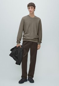 Brauner strukturierter Pullover, dunkelbraune Hose, hält eine schwarze Jacke, trägt schwarze Sportschuhe, steht vor einem hellgrauen Hintergrund.