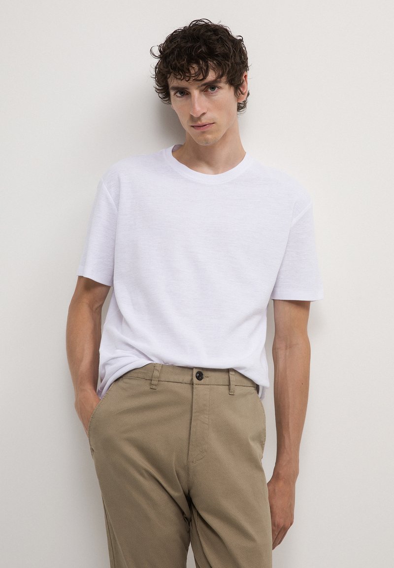 Uomo con capelli ricci che indossa una camicia bianca a maniche corte e pantaloni beige, in piedi contro un muro di colore chiaro.