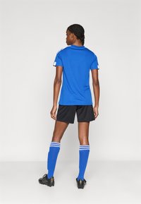 Μπλε αθλητική φανέλα με λευκές λεπτομέρειες, μαύρο σορτς και μπλε κάλτσες μέχρι το γόνατο με λευκές ρίγες, συνδυασμένα με μαύρα παπούτσια ποδοσφαίρου.