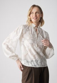 Blusa blanca con estampado floral, cuello redondo, detalle de cordón y puños elásticos, hecha de una tela ligera y semitransparente.