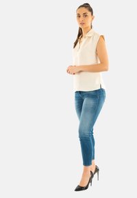 Blusa corta de manga corta en color crema con cuello, combinada con jeans ajustados azules y tacones altos de charol negros, con un diseño relajado y texturas sutiles.
