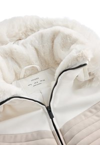 Bijela i bež pufasta jakna s umjetnom krznom, s patentnim zatvaračem i oznakom za njegu unutra. Glatka vanjština i mekana tekstura.