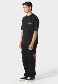 Jonge man in zwart T-shirt en zwarte cargobroek met rode grafische prints, zijwaarts staand tegen een eenvoudige lichte achtergrond.