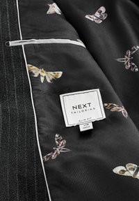 Slim-Fit-Sakko-Innenansicht mit schwarzem Stoff und Mottenmuster sowie Seitentasche. Etikett mit "NEXT TAILORING", Größe 40R.