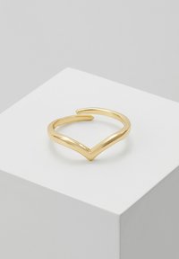 Bague en forme de V de ton or avec une finition lisse et polie. Le design est ouvert aux extrémités effilées, reposant sur un piédestal blanc.