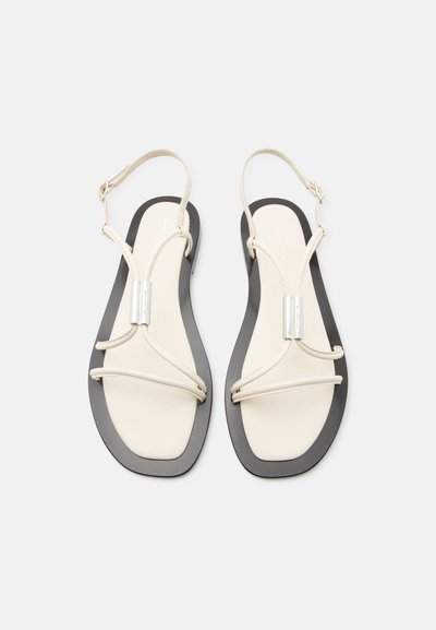 Μπεζ επίπεδα σανδάλια με λεπτά λουράκια, μεταλλικό ορθογώνιο στοιχείο, αγκράφες στον αστράγαλο και μαύρες άκρες στη σόλα σε λευκό φόντο.