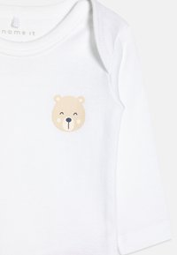 Body de algodão branco com mangas longas, apresentando uma impressão de cara de urso bege no peito. O decote tem um acabamento suave e arredondado.