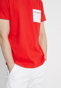 Červené tričko s krátkým rukávem a kulatým výstřihem, s bílou náprsní kapsou se značkovým logem "TOMMY JEANS". Měkký bavlněný materiál, volný střih.