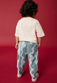 Világos krémszínű pamut póló, puha, kék, hullámmintás nadrággal párosítva, hátzsebekkel és fehér sportcipőkkel, vörös háttér előtt.