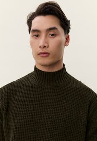 Fiatal férfi rövid sötét hajjal, sötétzöld, texturált kötött pulóvert visel egy egyszerű, világos háttér előtt.