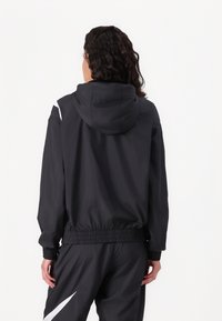 Svart huva-jacka med elastisk midjeresår och ärmslut. Har vita accenter på axlarna och en slät textur. Lättviktigt material.