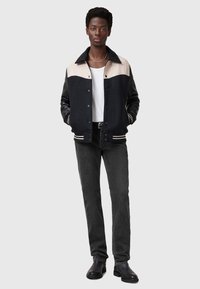 Musta ja kerma varsity-takki pyöreällä kauluksella, nappisuljenta ja raidalliset hihansuut, yhdistettynä tiukkoihin mustiin farkkuihin ja mustiin saappaisiin.