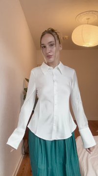 White button-down shirt with a collar, long sleeves, and a smooth texture, paired with a pleated teal skirt. Simple, elegant design.