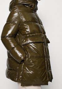 Chaqueta acolchada verde oliva, brillante, con capucha, que presenta un bolsillo lateral y una silueta cuadrada, textura acolchada y cierre de cremallera.