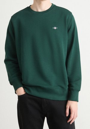 Person iført en mørkegrøn crewneck sweatshirt med en lille GANT-logo på venstre bryst, sorte bukser, den ene hånd i lommen.