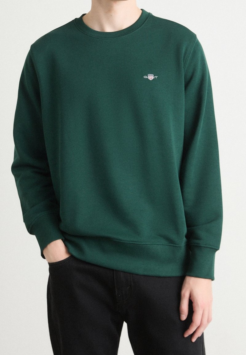 Personne portant un sweat-shirt vert foncé à col rond avec un petit logo GANT sur la poitrine gauche, pantalon noir, une main dans la poche.