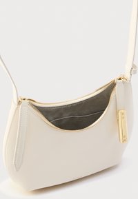 Ramenska torbica iz krem usnja s ukrivljeno obliko, zlato zadrgo in kovinskimi dodatki. Notranjost podložena s temno tkanino, opremljena z žepom z zadrgo.