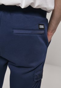 Marineblauwe joggingbroek met een geprofileerde afwerking, elastische taille, zijzakken en een klein zwart label met de tekst "URBAN CLASSICS" op de achterkant.