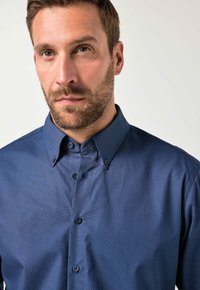 Hombre con cabello corto y marrón y barba, lleva una camisa de botones azul con un estampado, mirando ligeramente hacia un lado frente a un fondo claro y liso.