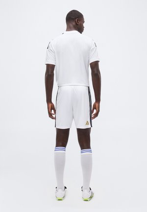 Hombre de espaldas con uniforme de fútbol blanco con rayas negras, calcetas blancas hasta la rodilla con rayas azules y botas blancas con suelas verdes.