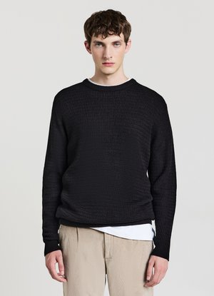 Giovane uomo rivolto verso l'obiettivo che indossa un maglione nero lavorato a maglia sopra una camicia bianca e pantaloni beige su uno sfondo chiaro e uniforme.