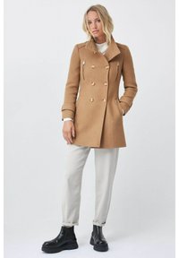 Abrigo tipo peacoat de mezcla de lana de color marrón claro con botones de doble botonadura, cremalleras laterales y un cuello con muesca, combinado con pantalones de color claro y botas de tobillo negras.