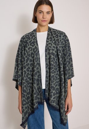 Kvinde iført et mørkegråt og sort leopardmønstret sjal over en hvid skjorte og blå jeans, stående mod en ensfarvet lys baggrund.