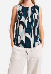 Ärmlös topp med abstrakt mönster i blått, svart och vitt, bärs med lösa vita byxor, händerna i fickorna.