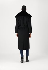 Palton negru cu un guler mare din blană artificială, talie cu brâu și mâneci lungi, combinat cu blugi închiși la culoare și cizme peste glezne.