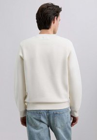 Sudadera blanca con cuello redondo, puños y dobladillo acanalados. Textura suave, acabado liso. Combinada con vaqueros azul claro, mostrando un estilo informal.