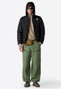 Chaqueta puffer negra, suéter de polar color topo, pantalones holgados verde oliva, botas marrones y accesorios que incluyen un bolso de cintura y gafas de sol.