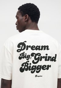 Mann mit kurzen lockigen Haaren, der ein weißes T-Shirt mit großem schwarzem Text „Dream Big, Grind Bigger“ auf dem Rücken trägt.