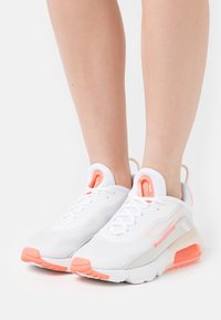 White athletic shoes with orange accents and a mesh upper. Features a rounded toe, laces, and cushioning in the midsole and heel.